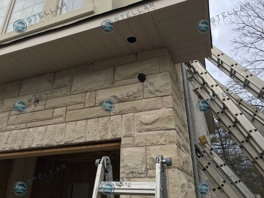 Dark security camera mounted on light wood-panel soffit above stone garage wall in Mississauga