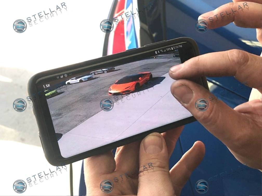 Smartphone screen displaying live remote security camera feed of exotic cars parked in auto shop lot.