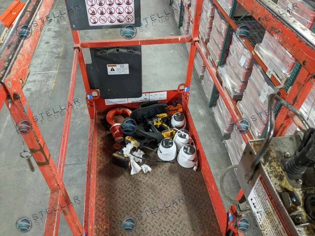 Scissor lift containing security camera equipment inside a large commercial warehouse during installation.