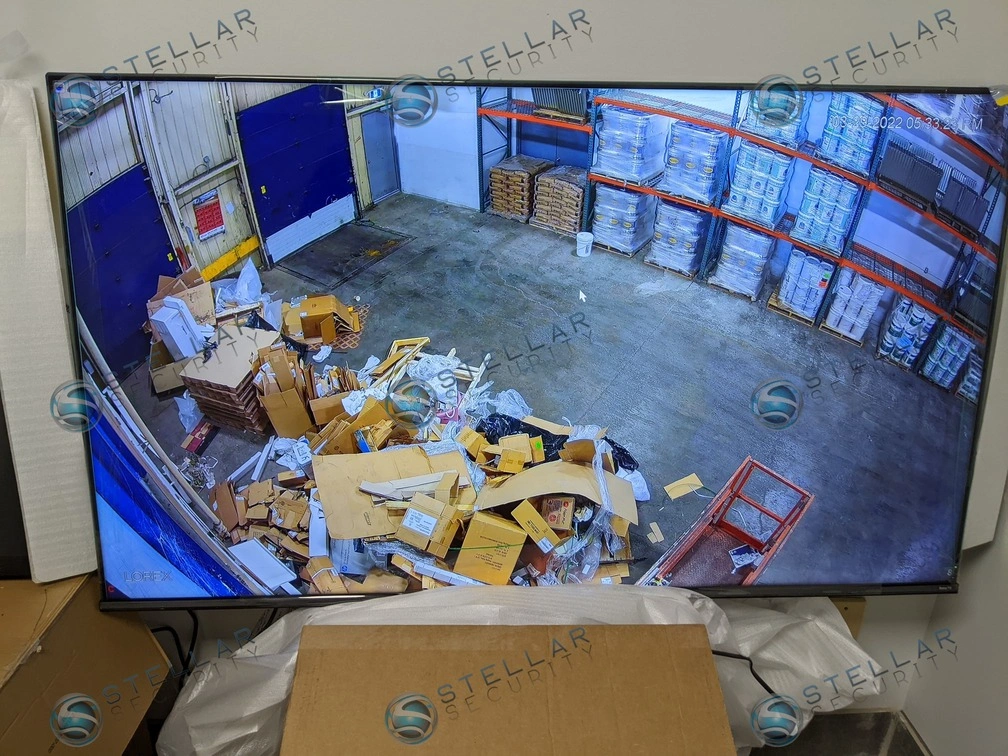 Wall-mounted TV monitor displaying real-time multi-channel security camera feeds of warehouse aisles and docks.