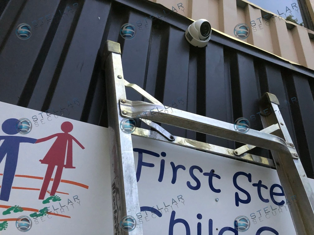White outdoor security camera mounted high above First Steps Childcare entrance signage on building exterior.