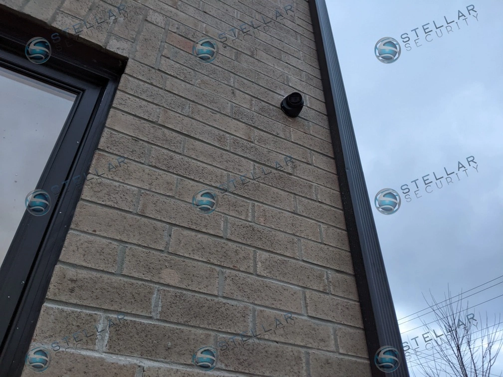 Black outdoor security camera mounted neatly on grey brick exterior wall near window frame in London
