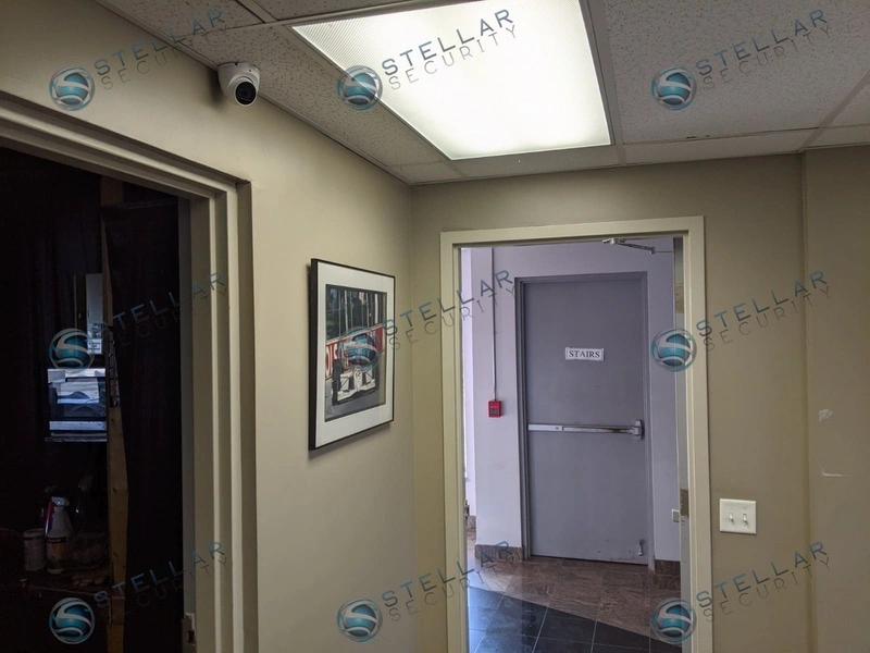 Compact dome security camera mounted on drop ceiling above office hallway and stairwell entrance.