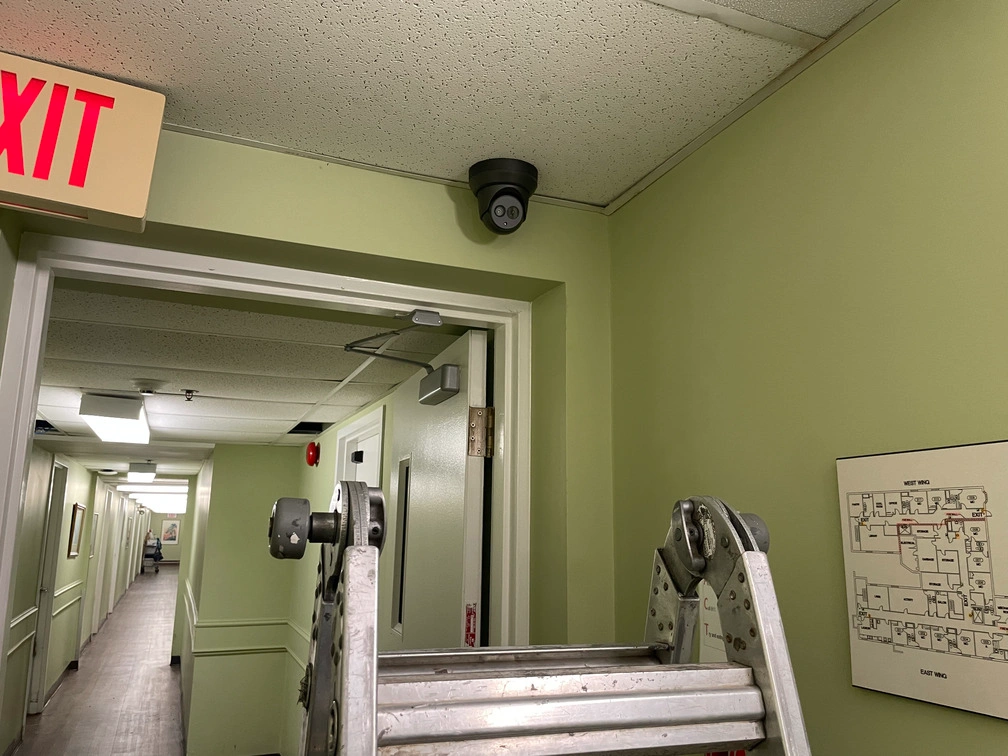High-angle wall view of black dome surveillance camera installed adjacent to exit sign and fire evacuation map.