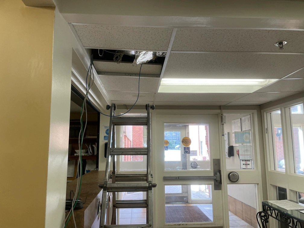 Ladder set up under ceiling tile access point near glass double door entrance inside retirement home vestibule.