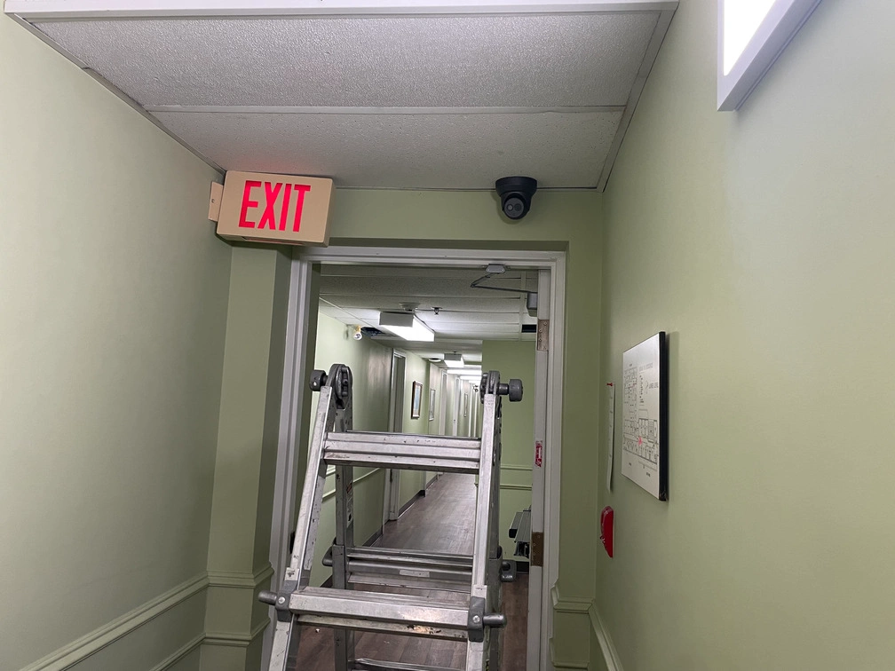 Stepladder placed in hallway doorway under illuminated exit sign during dome security camera ceiling installation.