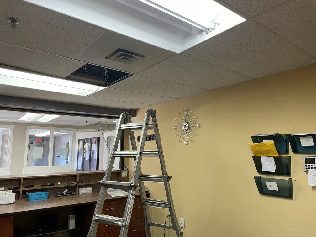 Stepladder positioned beneath opened drop ceiling tiles during security camera wiring at a retirement home reception desk.