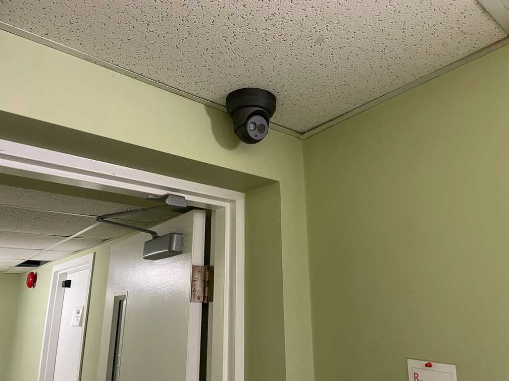 Close-up of black dome security camera mounted near ceiling corner above doorway in retirement facility hallway.