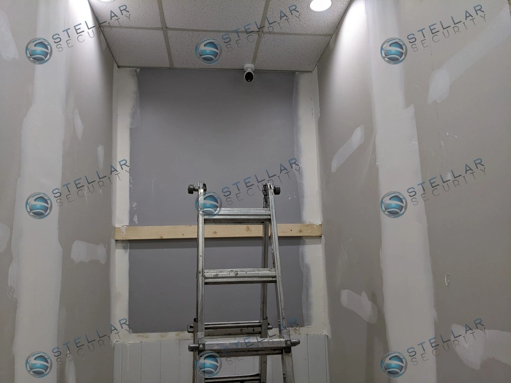 Step ladder positioned beneath a newly installed indoor security camera in a school hallway alcove.