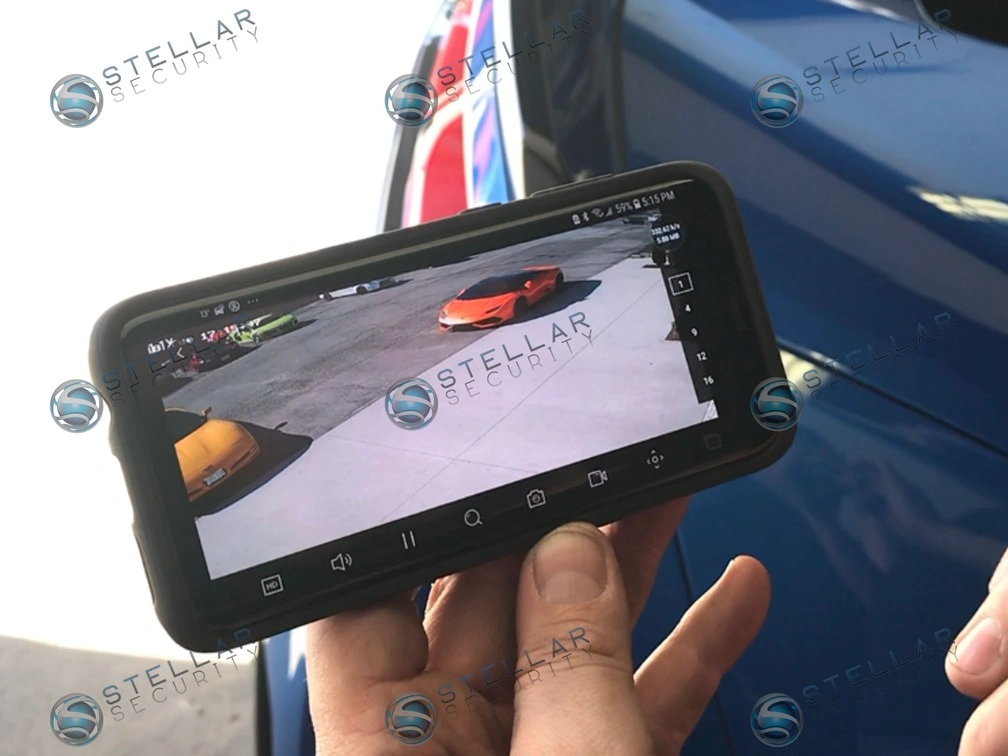 Technician holding smartphone displaying high-definition live security video feed of exotic sports cars.