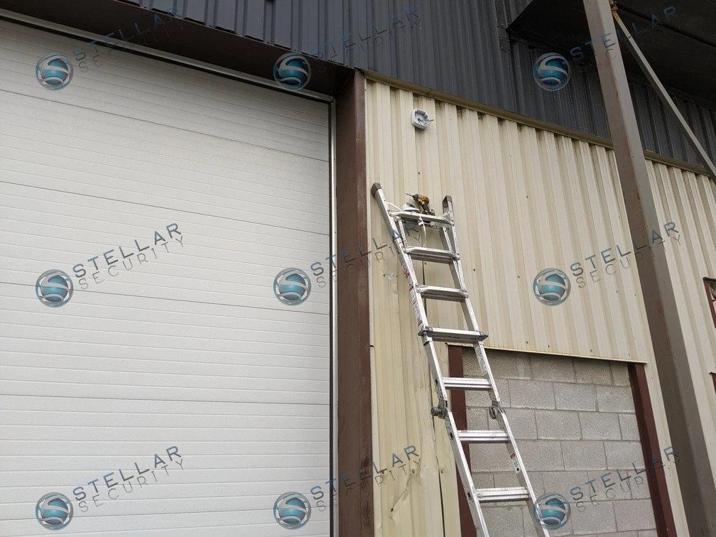 Exterior ladder setup against a metal warehouse wall for installing shipping bay door security cameras.